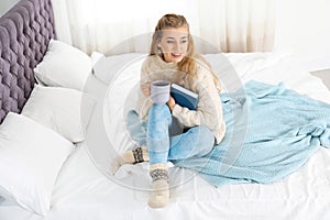 Attractive young woman in cozy warm sweater with cup of hot drink and book sitting on bed