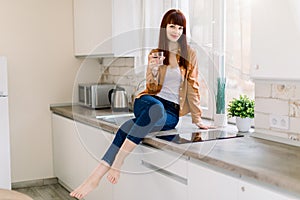 Attractive young woman in denim jeans and mustard casual shirt, sitting with glass of drinking water on the kitchen