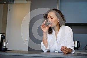 Attractive young woman dressed in white bathrobe, standing in the kitchen counter, drinking mineral water in the mornin