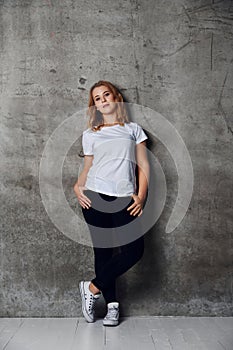 Attractive young woman dressed in a white blank t-shirt posing against a background of a concrete wall