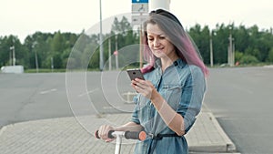 Attractive young woman with dyed hair in a denim dress and white sneakers riding a kick-scooter and using a smartphone.