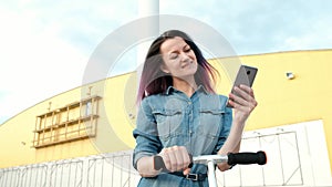 Attractive young woman with dyed hair in a denim dress and white sneakers riding a kick-scooter and using a smartphone.