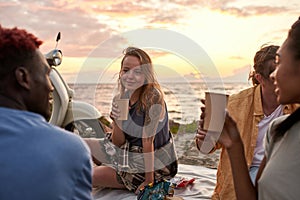 Attractive young woman enjoying picnic on the beach. Diverse friends having summer party at sunset