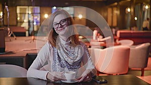 Attractive young woman with glasses enjoys coffee break relaxing in restaurant drinks coffee looks at camera and shows