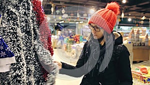 Attractive young woman in glasses and red hat chooses Christmas decor for a Christmas tree. The buyer found a cheap
