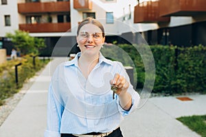 Attractive young woman in glasses showing key while standing outdoor against new house. Real estate agent holding key on