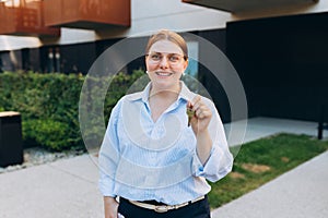 Attractive young woman in glasses showing key while standing outdoor against new house. Real estate agent holding key on