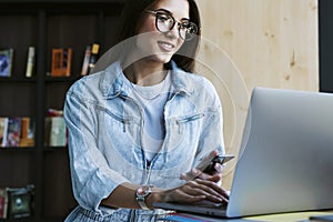 Attractive young woman in glasses stands near the window, holds smartphone in her hand and works on laptop.