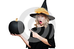 Attractive young woman in Halloween Witch costume holding large black pumpkin and pointing with a finger at it.