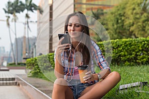 Attractive young woman in headphones listening to music using a smartphone and drinking coffee while sitting on curb