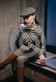 An attractive young woman in her 20s sits indoors, wearing a newsboy hat and stylish striped clothing, exuding elegance