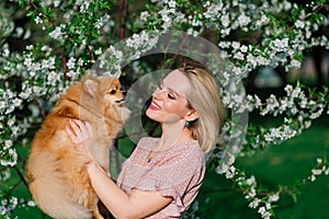 Attractive young woman holding dog spitz outside and smiling at camera, walking in the park