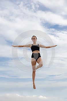 Attractive young woman jumping over cloudy blue sky. Caucasian woman wearing black sportswear. Fitness, wellness concept. Outdoor