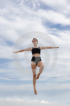 Attractive young woman jumping over cloudy blue sky. Caucasian woman wearing black sportswear. Fitness, wellness concept. Outdoor