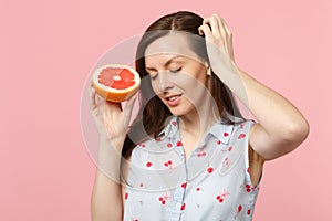 Attractive young woman keeping eyes closed put hand on hair holding half of fresh ripe grapefruit isolated on pink