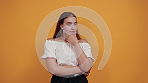 Attractive young woman keeping hand on chin, thinking about issue on orange wall