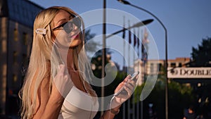 Attractive young woman listening to music in headphone use smartphone at city walk sunset look around smile portrait