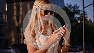 Attractive young woman listening to music in headphone use smartphone at city walk sunset look around smile portrait