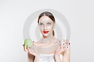 Attractive young woman looking at camera, holding in her hands green apple and glass of water promoting healthy lifestyle, diet