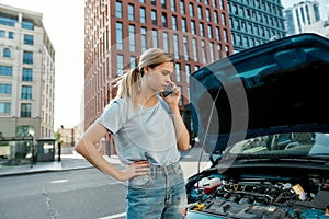 Attractive young woman looking dissatisfied, calling emergency car service, talking on the phone while standing near her