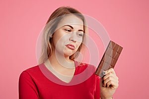 Attractive young woman looks with discontent expression at sweet bar of chocolate, keeps to diet, can`t eat it to be slim and spor