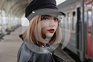 Attractive young woman millenial in black clothes and a hat and glasses at the railway station next to the train