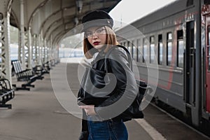 Attractive young woman millenial in black clothes and a hat and glasses at the railway station next to the train