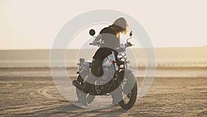 Attractive young woman motorcyclist makes a burnout on a motorcycle and ride away. Sunset or sunrise shot at desert. Female biker.