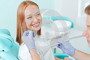 Attractive young woman with natural white teeth in dental clinic. Hands doctor dentist with teeth color palette next by face.