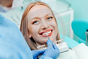 Attractive young woman with natural white teeth in dental clinic. Hands doctor dentist with teeth color palette next by face.