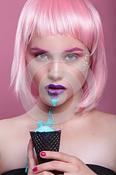 Attractive young woman with pink bob cut and purple lips eating blue ice cream on pink background