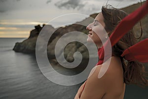 An attractive young woman poses in the summer against the background of the sea and mountains at sunset. Dressed in a red dress