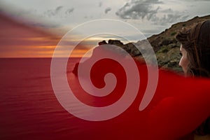 An attractive young woman poses in the summer against the background of the sea and mountains at sunset. Dressed in a red dress