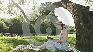 Attractive young woman reading book in park by the river. Brunette female sitting on meadow near tree in park and reading book dia