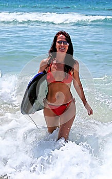 attractive young woman in red bikini walking in from blue sea on sunny beach with body board.