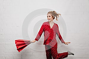 Attractive young woman in red cotton pajamas dancing with paper bag. Indoor portrait of happy blonde girl posing with