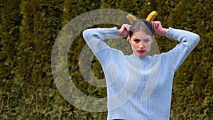 Attractive young woman with red lips in blue sweater puts bananas to her head as horns and imitates angelic imp