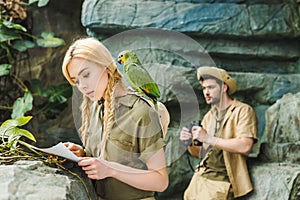 attractive young woman in safari suit with parrot and map navigating in jungle while her boyfriend