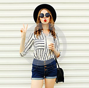 Attractive young woman sending sweet air kiss holding coffee cup in black round hat, shorts, white striped shirt on white wall