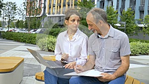 Attractive young woman and senior man are using laptop sitting on the bench in modern residential complex. Casual