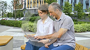 Attractive young woman and senior man are using laptop sitting on the bench in modern residential complex. Casual