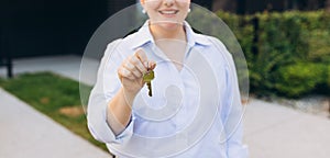 Attractive young woman showing key while standing outdoor against new house. Real estate agent holding key on street