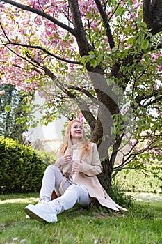 Attractive young woman sitting on lawn under blossoming tree, warm spring day. Vertical frame