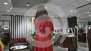 Attractive young woman is speaking on her smartphone dressed in red dress at clothing store, tracking shot, 360 degree