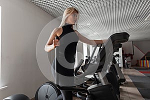 Attractive young woman in sporty black clothes on a stepper simulator in a gym. Girl is engaged in walking in fitness studio.