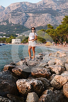 Attractive young woman stand on rocks by the sea. Stylish female in front of big mountains