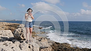 Attractive young woman standing on rock at seashore and looking to the phone. Caucasian brunette girl standing near sea with phone