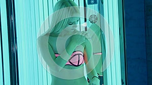 Attractive young woman standing in tanning booth and applying a cream on body.