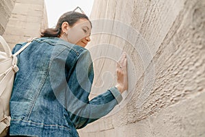 Attractive young woman stands between concrete walls. Caucasian girl in casual jeans and dress looks back