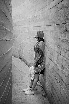 Attractive young woman stands between concrete walls. Caucasian girl in casual jeans and flower dress looks up.